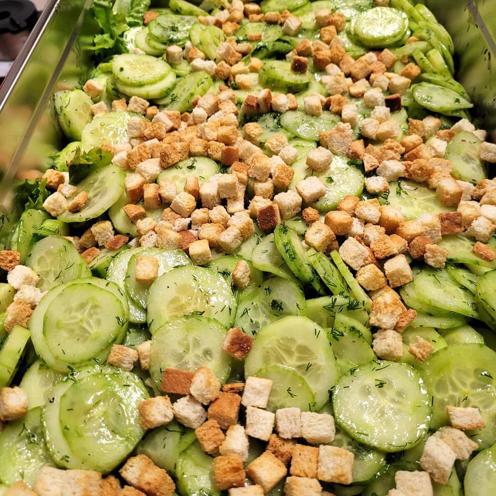 Frischer Gurkensalat mit Dill und Croutons auf grünem Salatblattbett in einem silbernen Tablett.