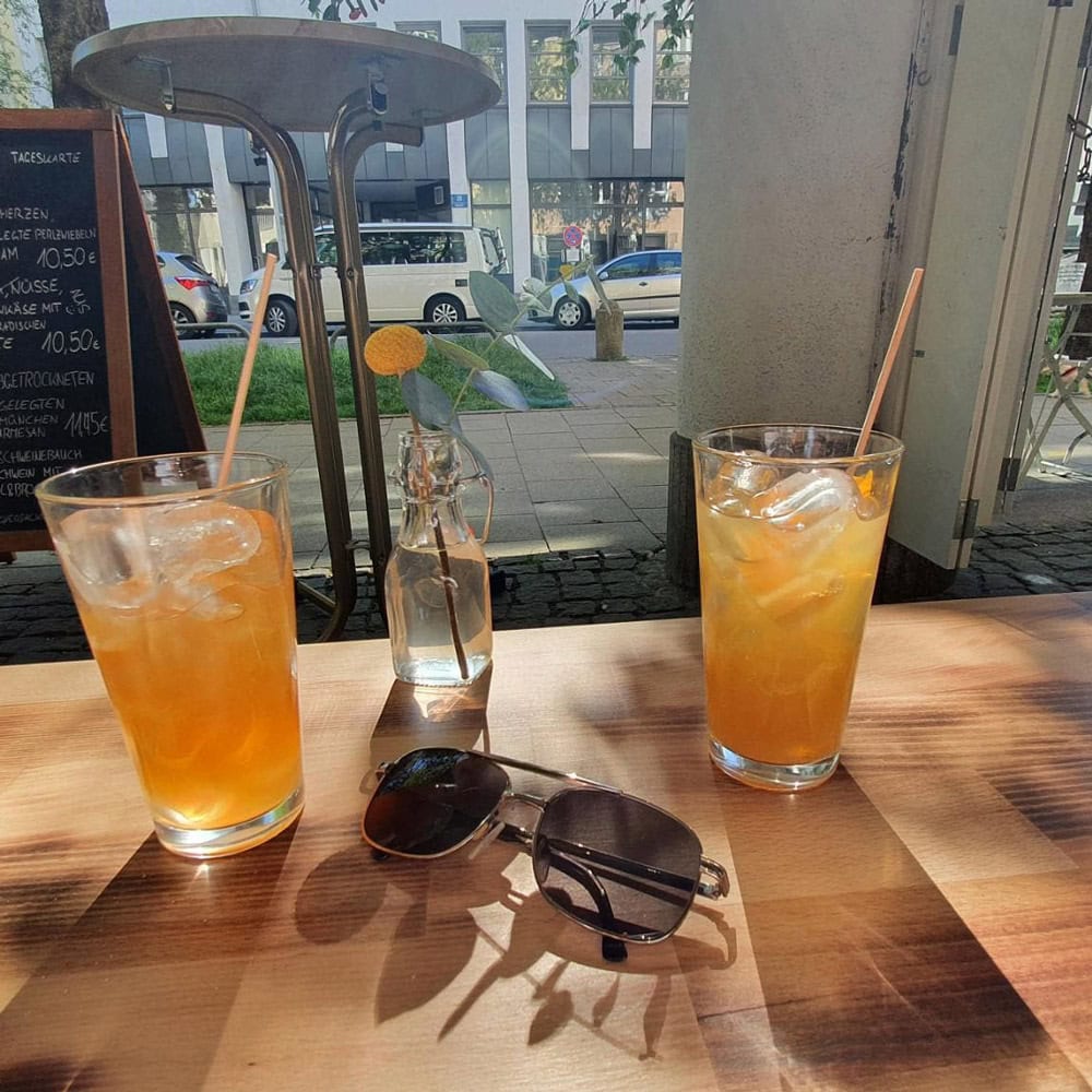 Zwei Gläser mit Eistee und Strohhalmen auf einem Holztisch, Sonnenbrille, sonnige Straßenszene.